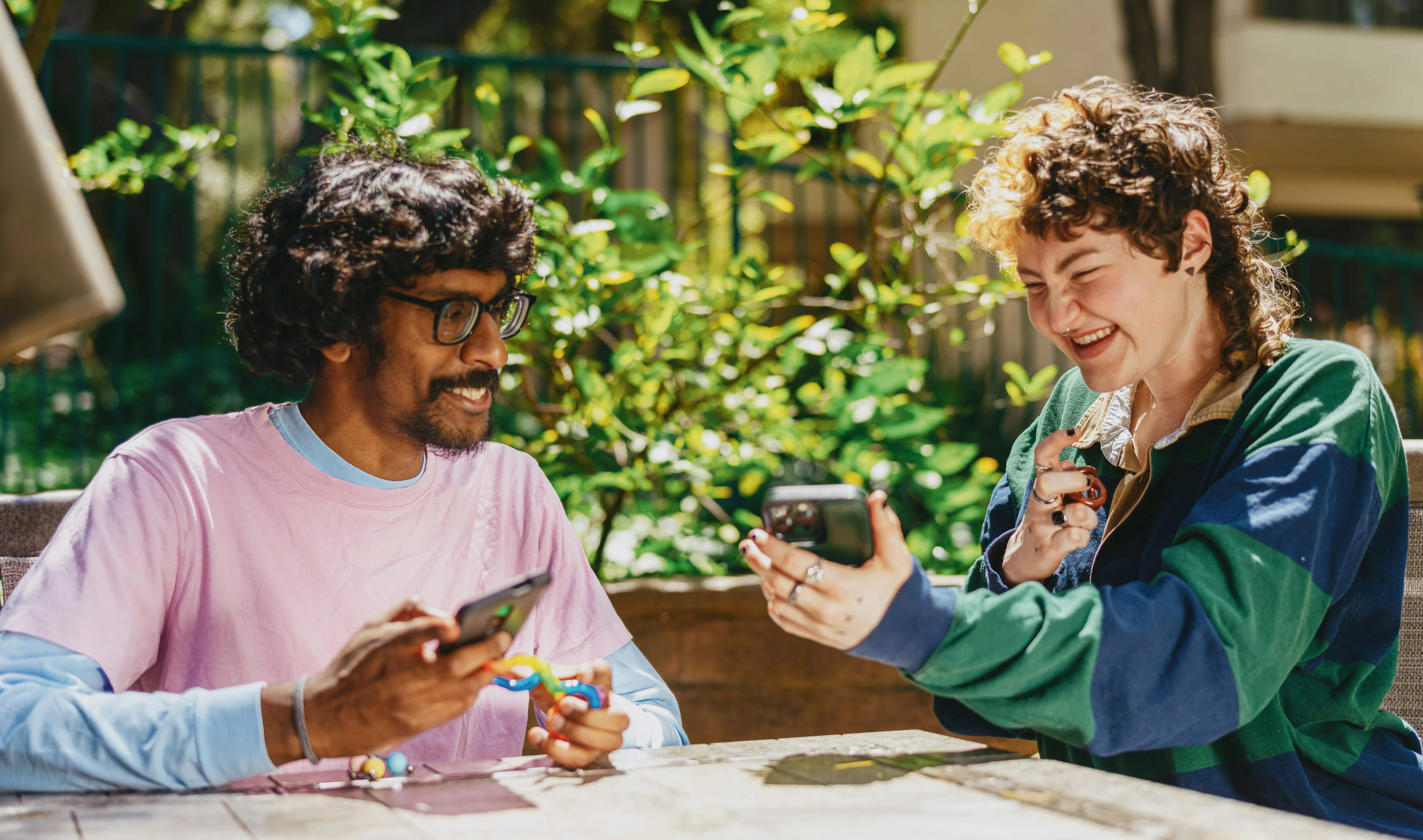 Two people outdoors laugh together while looking at a phone; one holds a fidget toy.